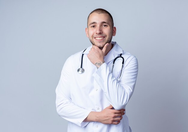 Jovem médico sorridente, vestindo bata médica e estetoscópio em volta do pescoço, tocando seu queixo isolado na parede branca