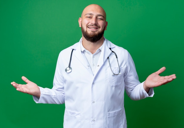 Foto grátis jovem médico sorridente, vestindo bata médica e estetoscópio espalhando as mãos isoladas no verde