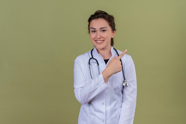 Jovem médico sorridente, vestindo jaleco médico e estetoscópio apontando para o lado na parede verde