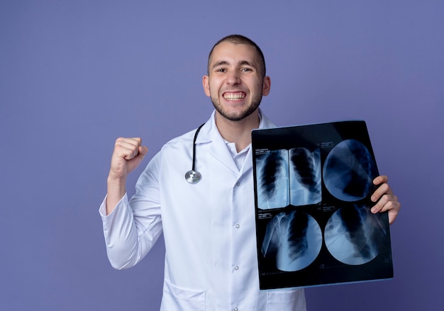 Foto grátis jovem médico sorridente, vestindo túnica médica e estetoscópio em volta do pescoço, segurando uma foto de raio-x e cerrando o punho isolado na parede roxa