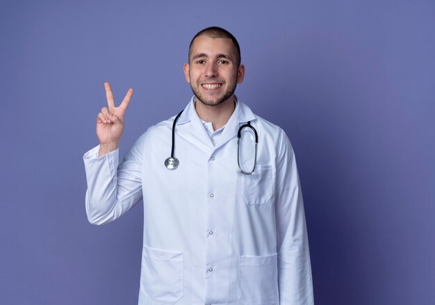 Jovem médico sorridente, vestindo túnica médica e estetoscópio, fazendo o sinal da paz isolado na parede roxa