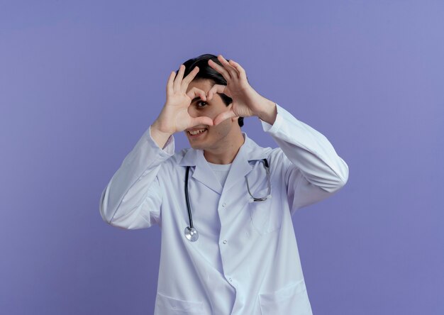 Foto grátis jovem médico sorridente, vestindo túnica médica e estetoscópio fazendo um sinal de coração, olhando através dele isolado