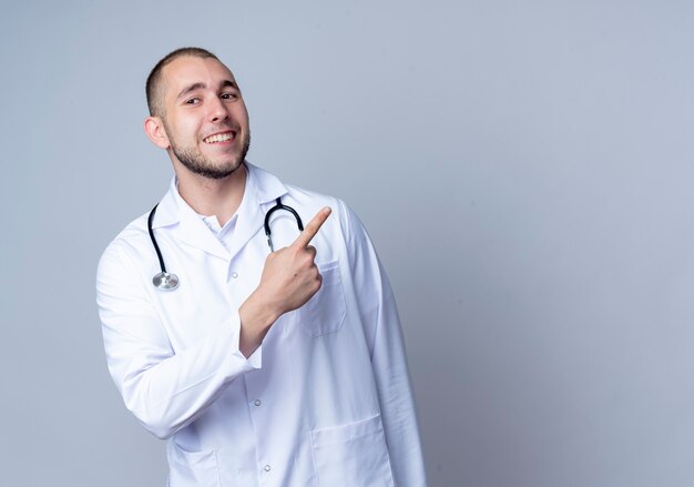 Jovem médico sorridente, vestindo túnica médica e estetoscópio no pescoço, apontando para o lado isolado na parede branca