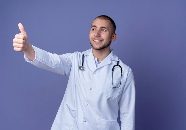 Jovem médico sorridente, vestindo túnica médica e estetoscópio no pescoço, olhando para o lado e mostrando o polegar isolado na parede roxa