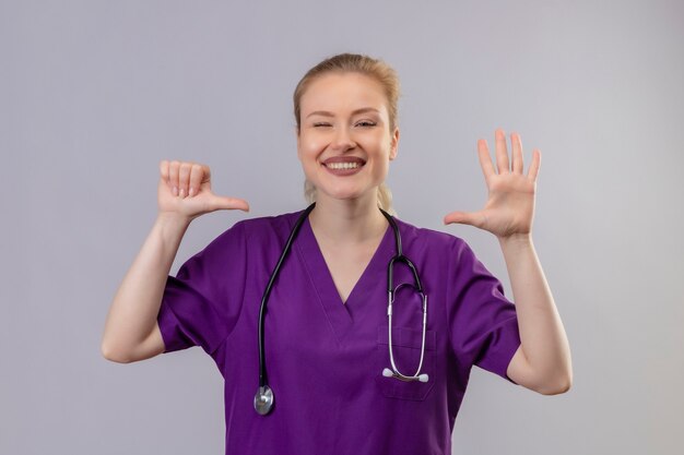 Jovem médico sorridente, vestindo um jaleco roxo e um estetoscópio mostrando diferentes gestos na parede branca isolada