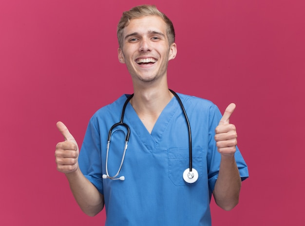 Jovem médico sorridente, vestindo uniforme de médico com estetoscópio aparecendo os polegares isolados na parede rosa