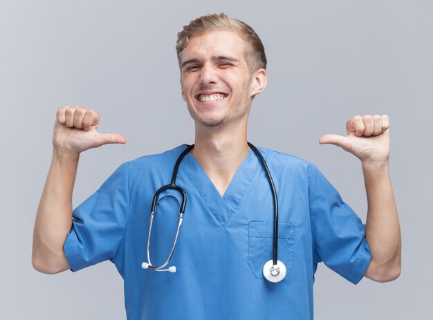 Jovem médico sorridente, vestindo uniforme de médico com estetoscópio apontando para si mesmo, isolado na parede branca