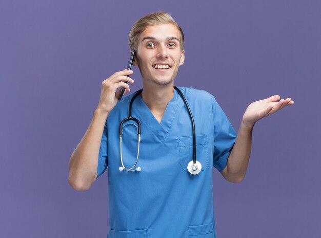 Jovem médico sorridente, vestindo uniforme de médico com estetoscópio, falando no telefone, espalhando a mão isolada na parede azul