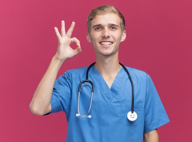 Jovem médico sorridente, vestindo uniforme de médico com estetoscópio, mostrando um gesto de aprovação isolado na parede rosa