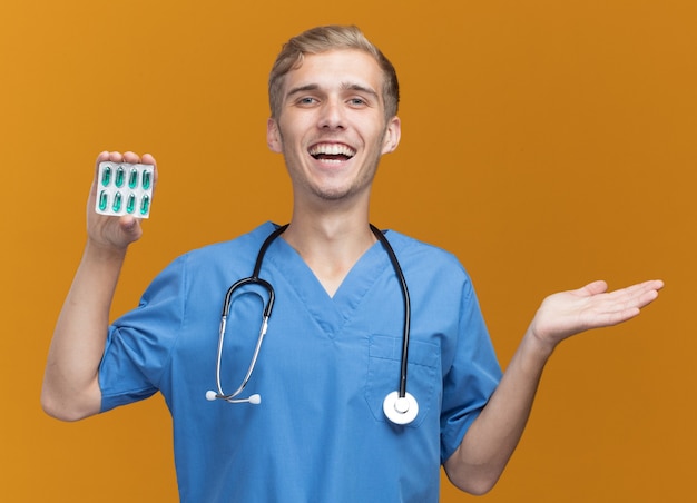 Jovem médico sorridente, vestindo uniforme de médico com estetoscópio segurando comprimidos e espalhando a mão isolada na parede laranja