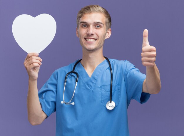 Jovem médico sorridente, vestindo uniforme de médico com estetoscópio segurando uma caixa em forma de coração e mostrando o polegar isolado na parede azul
