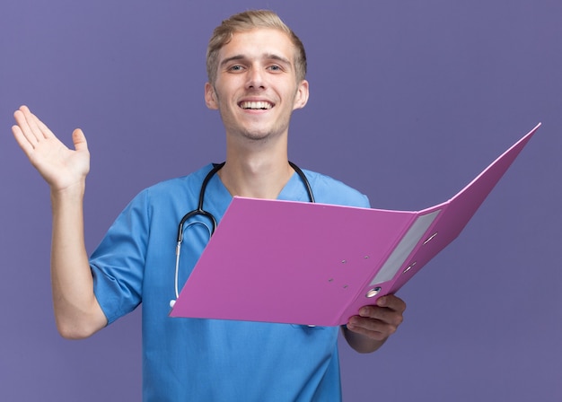 Foto grátis jovem médico sorridente vestindo uniforme de médico com estetoscópio segurando uma pasta e espalhando a mão isolada na parede azul