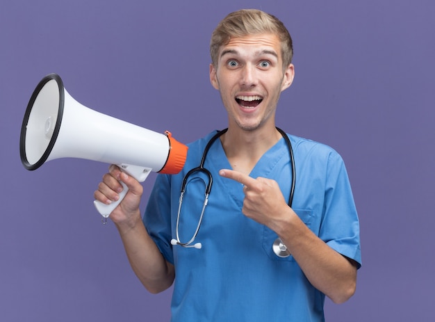 Jovem médico sorridente, vestindo uniforme de médico, segurando um estetoscópio e apontando para um alto-falante isolado na parede azul