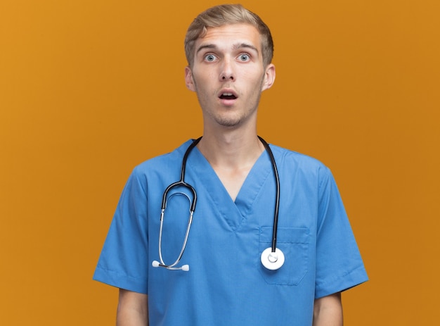 Jovem médico surpreso usando uniforme de médico com estetoscópio isolado na parede laranja