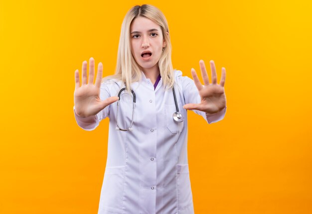 Jovem médico triste usando estetoscópio e vestido de médico, mostrando um gesto de parada com as duas mãos sobre fundo amarelo isolado