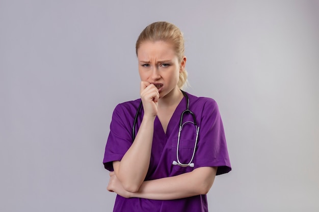 Jovem médico triste vestindo um vestido médico roxo e estetoscópio segurando o queixo na parede branca isolada