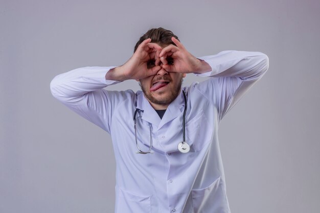 Jovem médico vestindo jaleco branco e estetoscópio fazendo okey gesto como binóculos furando a língua de fora, olhando por entre os dedos, expressão maluca