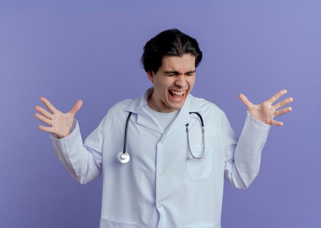 Jovem médico zangado usando túnica médica e estetoscópio, mostrando as mãos vazias, gritando com os olhos fechados, isolados na parede roxa