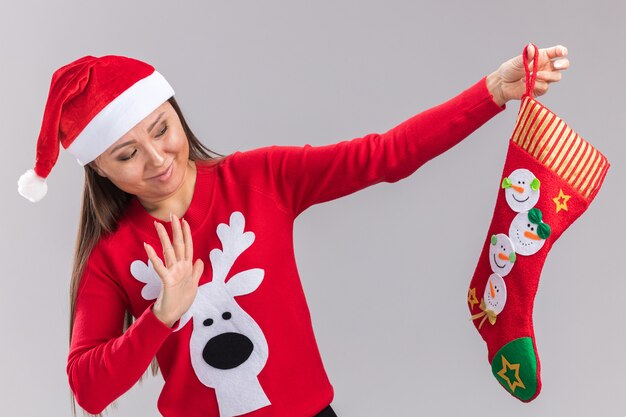 Jovem menina asiática com chapéu de natal e suéter, segurando e olhando para uma meia de natal isolada no fundo branco