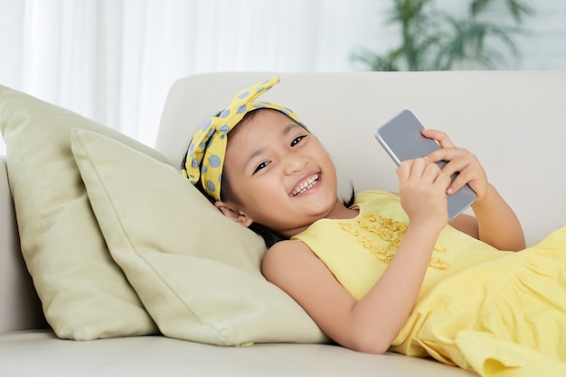 Jovem menina asiática, deitado no sofá com o smartphone, olhando para a câmera e sorrindo