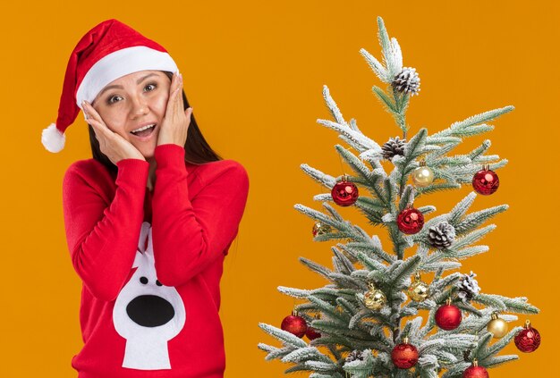 Jovem menina asiática sorridente com chapéu de natal e suéter em pé perto da árvore de natal, colocando as mãos nas bochechas isoladas em fundo laranja