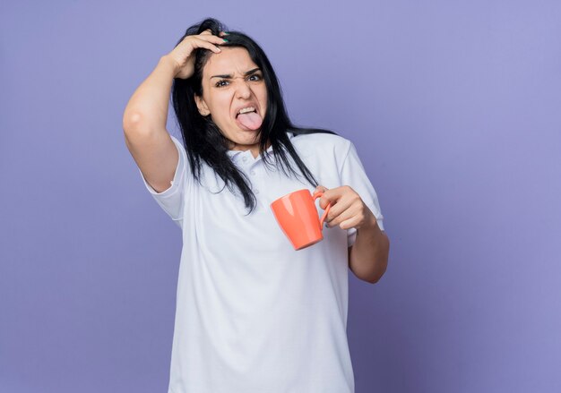 Jovem menina morena caucasiana irritada mostra a língua e levanta o cabelo segurando um copo isolado na parede roxa