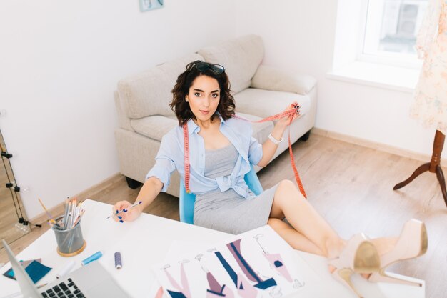 Jovem menina morena com um vestido cinza e uma camisa azul se senta em uma mesa na oficina. Ela colocou suas longas pernas com saltos na mesa. Ela segura o cinto na mão.