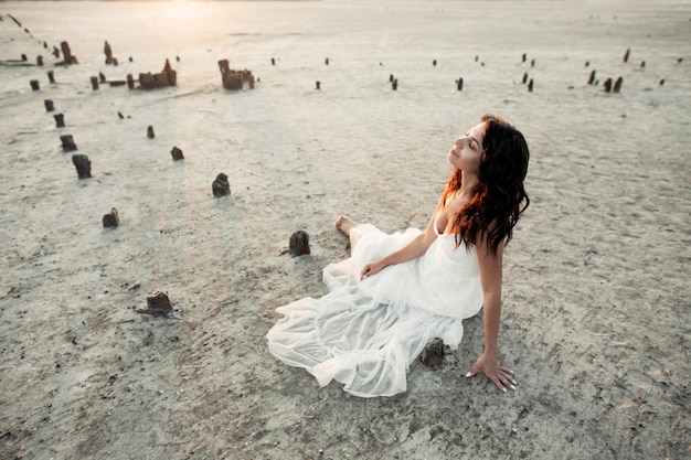 Jovem menina morena está sentado na areia com os olhos fechados, vestida de branco casual vestido