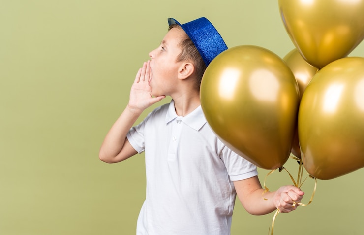 Foto grátis jovem menino eslavo com chapéu de festa azul segurando balões de hélio e mantendo a mão perto da boca chamando alguém olhando para o lado isolado na parede verde oliva com espaço de cópia