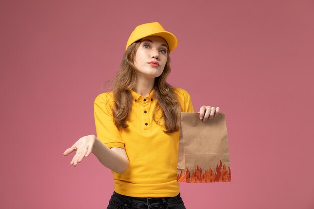 Foto grátis jovem mensageira com capa amarela e uniforme amarelo segurando um pacote de entrega de comida na cor de serviço de entrega uniforme de fundo rosa escuro