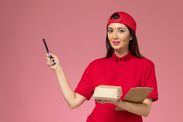 Jovem mensageira com capa de uniforme vermelha e um pequeno pacote de comida para entrega e um bloco de notas com uma caneta nas mãos na parede rosa claro.