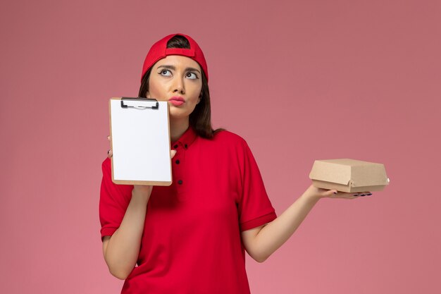 Jovem mensageira com capa de uniforme vermelha e um pequeno pacote de comida para entrega e um bloco de notas nas mãos, pensando na parede rosa