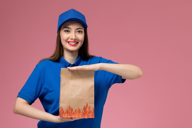 Jovem mensageira de frente com uniforme azul e capa segurando um pacote de comida de papel e sorrindo na mesa rosa claro