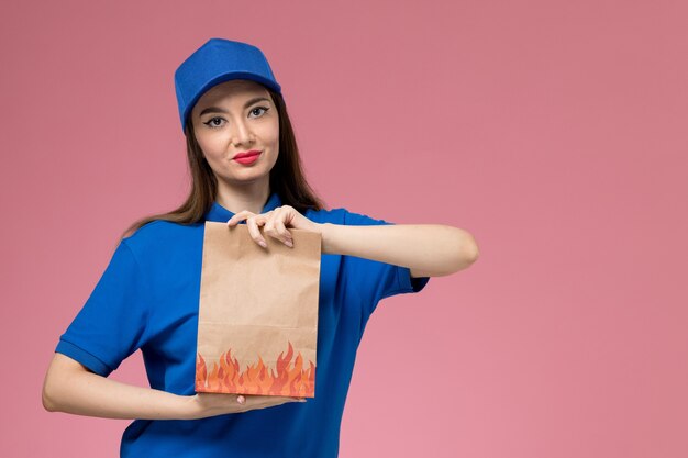 Jovem mensageira de frente com uniforme azul e capa segurando um pacote de comida na parede rosa claro.