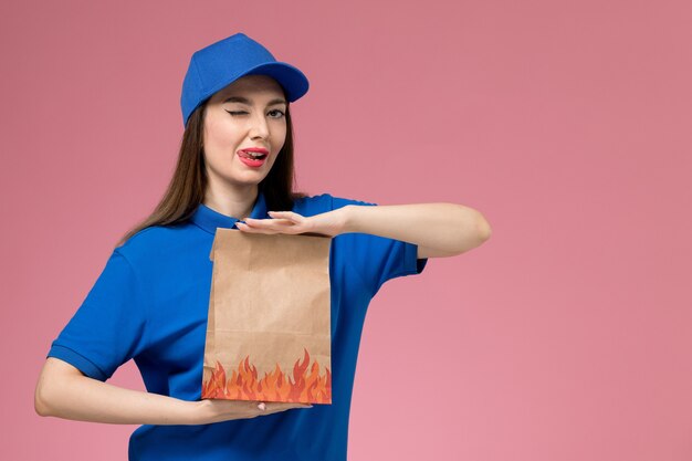 Jovem mensageira de frente com uniforme azul e capa segurando um pacote de comida piscando na parede rosa claro