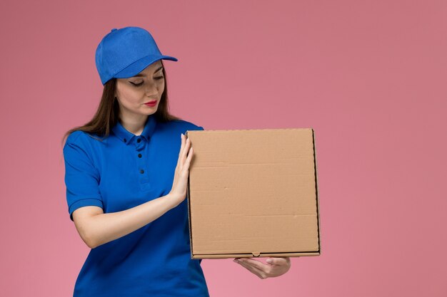 Jovem mensageira de frente com uniforme azul e capa segurando uma caixa de comida na parede