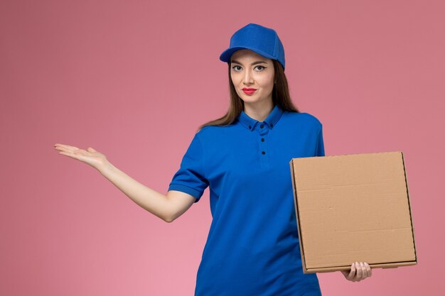 Jovem mensageira de frente com uniforme azul e capa segurando uma caixa de entrega de comida na parede rosa woorker