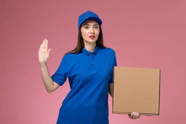 Jovem mensageira de frente com uniforme azul e capa segurando uma caixa de entrega de comida na parede rosa