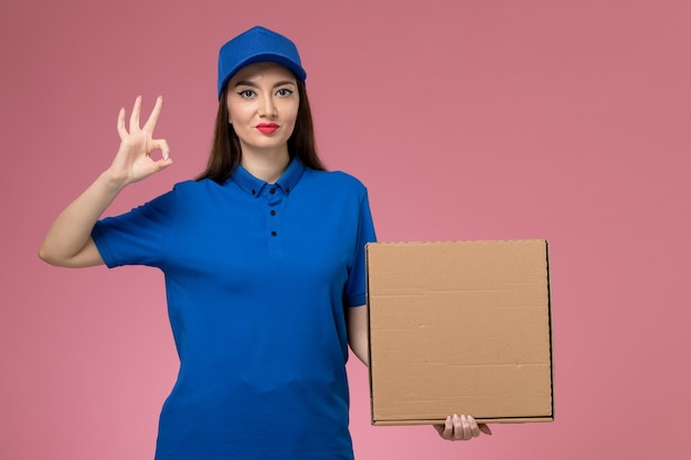 Jovem mensageira de frente com uniforme azul e capa segurando uma caixa de entrega de comida na parede rosa