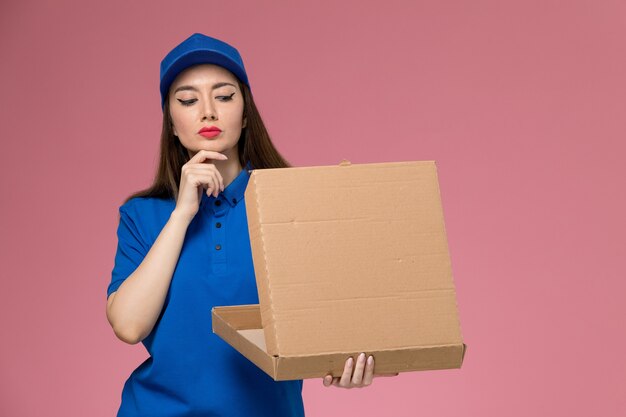 Jovem mensageira de frente com uniforme azul e capa segurando uma caixa de entrega de comida pensando na parede rosa