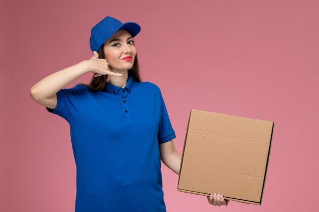 Jovem mensageira de frente com uniforme azul e capa segurando uma caixa de entrega de comida, posando na parede rosa