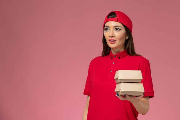 Jovem mensageira de frente com uniforme vermelho e capa com pequenos pacotes de entrega de comida nas mãos na parede rosa