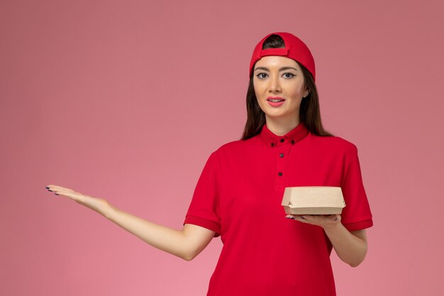Jovem mensageira de frente com uniforme vermelho e capa com um pequeno pacote de entrega de comida nas mãos na parede rosa