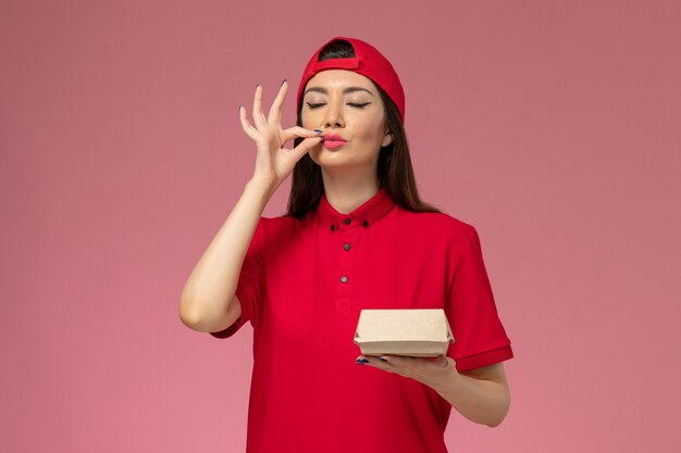 Jovem mensageira de frente com uniforme vermelho e capa com um pequeno pacote de entrega de comida nas mãos na parede rosa