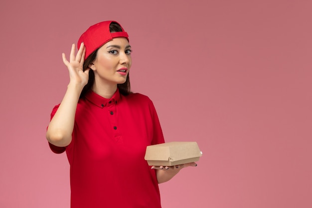 Jovem mensageira de frente com uniforme vermelho e capa com um pequeno pacote de entrega de comida nas mãos na parede rosa