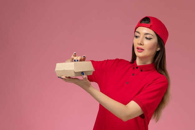 Jovem mensageira de frente com uniforme vermelho e capa com um pequeno pacote de entrega de comida nas mãos na parede rosa