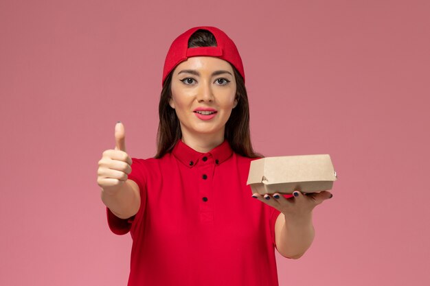 Jovem mensageira de frente com uniforme vermelho e capa com um pequeno pacote de entrega de comida nas mãos na parede rosa