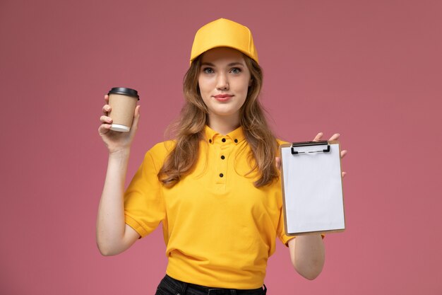 Jovem mensageira de uniforme amarelo segurando a xícara de café e o bloco de notas na mesa rosa escuro. trabalho de entrega de uniforme.