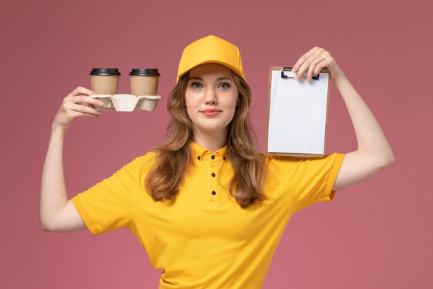 Jovem mensageira de uniforme amarelo segurando copos de café de plástico e um bloco de notas na mesa rosa escuro. Trabalhador de serviço de entrega de uniforme