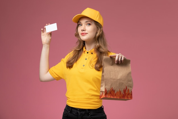 Foto grátis jovem mensageira de uniforme amarelo segurando um cartão branco de pacote de comida de entrega no fundo rosa.